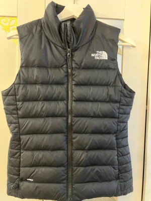 Svart dunväst från The North Face - Snygg svart dunväst från The North Face med hög krage och dragkedja framtill. Västen har två sidofickor och diskret logga på bröstet och ryggen. Perfekt för lager-på-lager och håller dig varm under kyliga dagar. Klassisk quiltad design i lätt och smidigt material.