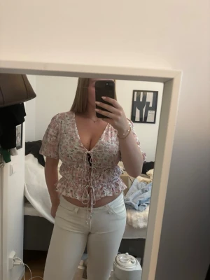 Blommig blus - Söt blommig blus från AWARE i vitt med rosa och grått blommönster. Modellen har djup urringning, korta puffärmar och knytning framtill.