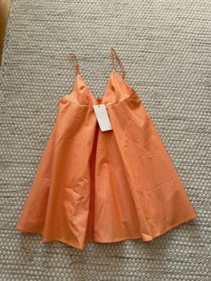  Ny trendig Orange  sommarklänning Zara  - Säljer en luftig kort klänning med tunna axelband i orange och vitt randigt mönster. Klänningen har en A-linjeformad siluett och är perfekt för varma dagar. Materialet känns lätt och bekvämt mot huden.