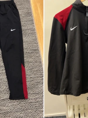 Nike FC Barcelona träningsset svart/röd - Säljer ett svart träningsset från Nike med FC Barcelona-logga. Tröjan har dragkedja, röd och blå detalj på axlarna samt vit Nike-logga. Byxorna är svarta med röd detalj och vit Nike-logga. Perfekt för fotbollsträning eller gymmet.