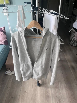 Ljusgrå hoodie med dragkedja - Snygg ljusgrå hoodie från Polo Ralph Lauren med dragkedja framtill och två fickor. Klassisk huva och liten broderad logga på bröstet. Tillverkad i mjukt bomullsmaterial som känns skönt mot huden. Perfekt för en avslappnad och stilren look.