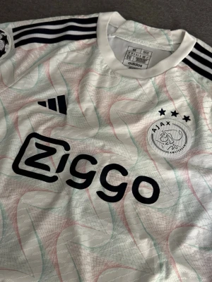 Ajax Authentic Tröja - Snygg Ajax matchtröja från adidas i vitt med svarta detaljer och diskreta mönster i grönt och rött. Tröjan har Ziggo-logga på bröstet, Ajax klubbmärke med tre stjärnor och svarta ränder på axlarna. Perfekt för fotbollsträning eller att bära till vardags.