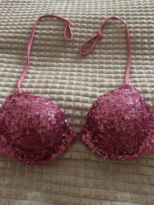 Rosa paljett-bh med halterneck - Supersnygg rosa bh täckt med glittriga paljetter och tunna halterneckband som knyts i nacken. Kuporna är vadderade och ger en push-up effekt. Perfekt för dig som vill sticka ut och glänsa lite extra.