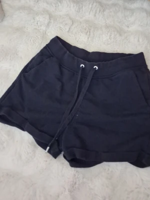 Svarta mjukisshorts med snörning - Ett par svarta mjukisshorts med bred resår i midjan och snörning framtill. Shortsen har två sidofickor och är tillverkade i ett mjukt bomullsmaterial. Perfekta för chill dagar eller träning.