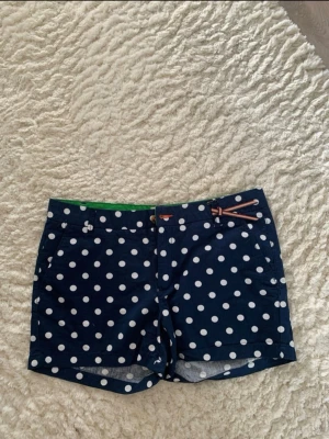 Marinblå prickiga shorts  - Snygga marinblå shorts med vita prickar. Shortsen har fickor fram och detaljer i grönt på insidan av linningen. Perfekta för varma dagar och ger en lekfull touch till din outfit. Köpta från vinted men har inte andvända denna sommaren så därför säljer jag dem. 