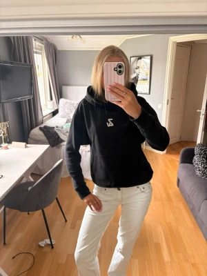 Svart hoodie med tryck från Zadig & Voltaire - Säljer en svart hoodie från Zadig & Voltaire med vit logga framtill och stort grafiskt tryck på ryggen med texten 'GLAMROCK FESTIVAL' och en bild på en telefonlur. Hoodien har huva och magficka. Perfekt för dig som gillar streetwear och coola detaljer.