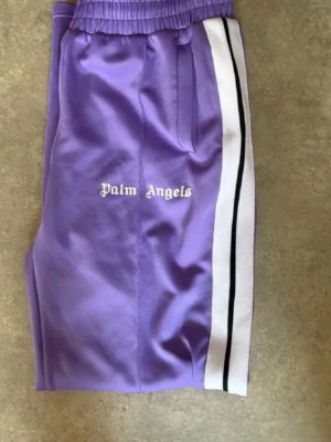 Lila byxor från Palm Angels - Säljer ett par lila byxor från Palm Angels med vit och svart rand längs sidorna. Byxorna har elastisk midja och fickor, samt Palm Angels-logga broderad framtill. Perfekta för en avslappnad och trendig look.