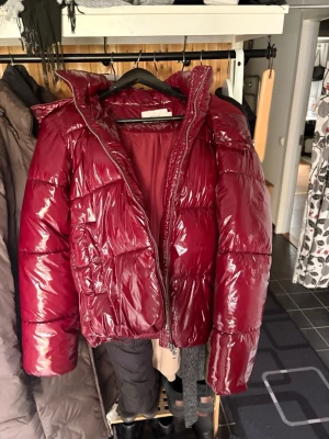 Vinröd pufferjacka från Nelly - Snygg vinröd pufferjacka från Nelly med glansig finish och vadderad design. Jackan har dragkedja framtill, hög krage och stora puffiga ärmar. Perfekt för kalla dagar när du vill hålla stilen och värmen. Dock är det ett litet hål på ena ärmen som säkert går att fixa om man kan sånt.😊💕