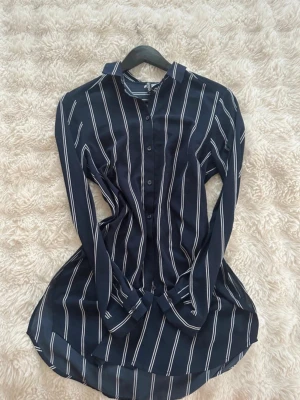 Mörkblå randig skjorta med knappar - Säljer en långärmad skjorta i mörkblått med vita vertikala ränder. Skjortan har klassisk krage, knäppning framtill och rundad nederkant. Perfekt till jeans eller kjol för en chill och stilren look.