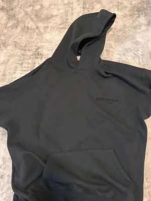 Svart Essentials Fear of God hoodie - Säljer en svart hoodie från Essentials Fear of God med broderad logga på bröstet och stor text på ryggen. Hoodien har huva, magficka och ribbade muddar. Tillverkad i mjukt bomullsmaterial som är riktigt skönt att ha på sig. STORLEK M MEN PASSAR OVERSIZE