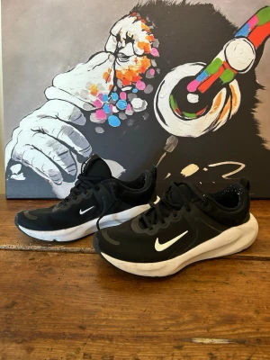 Svarta Nike sneakers med vit sula - Snygga svarta Nike sneakers med vit sula och vit swoosh på sidan. Skorna har en sportig design med rundad tå, svarta skosnören och detaljer i mesh för extra andningsförmåga. Yttersulan har vågigt mönster och synlig dämpning i hälen.