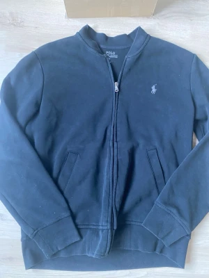Svart zip hoodie från Polo Ralph Lauren - Klassisk svart hoodie med dragkedja från Polo Ralph Lauren. Tröjan har två fickor framtill, ribbade muddar och den ikoniska loggan broderad på bröstet. Tillverkad i mjukt bomullsmaterial och har en stilren, enkel design utan huva.