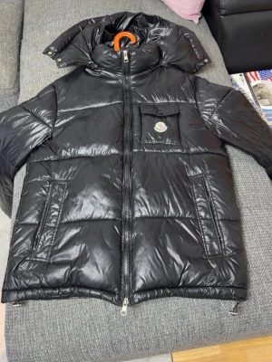 Svart dunjacka från Moncler - Snygg svart dunjacka från Moncler med glansig finish, stor huva och dragkedja framtill. Jackan har två sidofickor och en bröstficka med lock och Moncler-logga. Perfekt för kalla vinterdagar och riktigt trendig look.