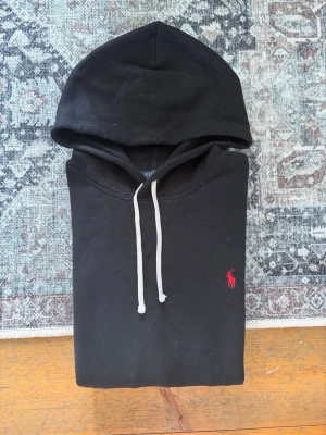 Svart hoodie med röd logga - Svart hoodie från Polo Ralph Lauren med klassisk röd broderad logga på bröstet. Hoodien har vit dragsko i huvan och en stor magficka framtill. Tillverkad i mjukt bomullsmaterial och har ribbade muddar vid ärmslut och nederkant.