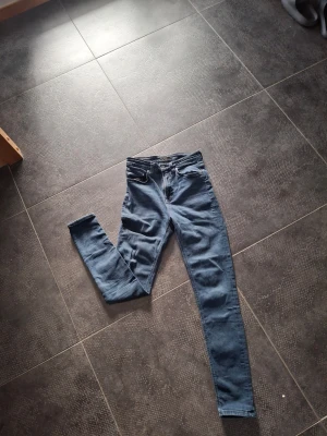 Nudie jeans - Säljer ett par mörkblå skinny jeans med klassisk femficksdesign och hög midja. Jeansen har dragkedjegylf och silverfärgade nitar. Materialet är stretchigt denim som sitter tajt längs benen.
