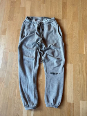 Grå Essentials mjukisbyxor - Snygga grå mjukisbyxor från Essentials med resår i midjan och benslut samt dragsko. Byxorna har två sidofickor och en avslappnad passform. Perfekta för chill dagar och streetwear-stil.