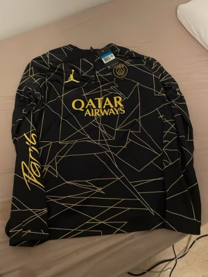 Svart PSG långärmad träningströja - Snygg svart långärmad träningströja från Paris Saint-Germain med guldiga detaljer och tryck. Tröjan har grafiskt mönster, PSG-logga på bröstet, Jordan-logga och Qatar Airways-tryck. Dragkedja vid halsen och Paris-tryck på ärmen.