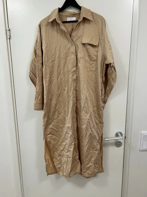 Beige skjortklänning från A Day's March - Stilren beige skjortklänning från A Day's March i storlek 38. Klänningen har lång ärm, klassisk krage, bröstficka och knäppning framtill. Tillverkad i ett mjukt, lätt material som ger en avslappnad look.