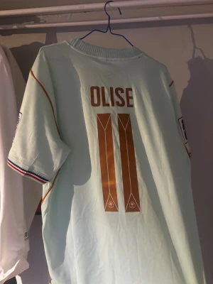 Frankrike fotbollströja Olise #11 - Ljusblå fotbollströja från Nike med Olise och nummer 11 på ryggen. Tröjan har Frankrikes landslagsemblem med två stjärnor, guldiga detaljer och randiga muddar i blått, vitt och rött. Korta ärmar och broderad Nike-logga på bröstet.