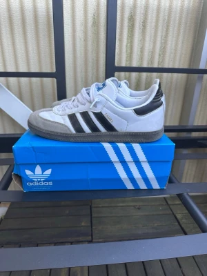 Adidas Samba OG vita och svarta sneakers - Klassiska Adidas Samba OG sneakers i vitt och svart med grå mockadetaljer och de ikoniska tre svarta ränderna på sidan. Låg modell med gummisula och ovandel i läder och mocka. Perfekta för en stilren och sportig look.