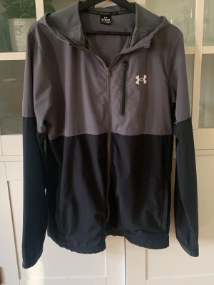 Under Armour windbreaker  - Snygg vindjacka från Under Armour i grått och svart med hel dragkedja, huva och en bröstficka med dragkedja. Jackan har lång ärm och logga på bröstet. Perfekt för blåsiga dagar och sportiga outfits.