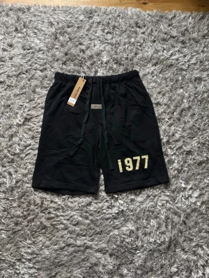 Svarta shorts Essentials 1977 - Svarta shorts från Essentials med trycket '1977' i vitt på vänster ben. Shortsen har snörning i midjan och en relaxed passform. Tillverkade i mjukt bomullsmaterial, perfekta för sommaren.