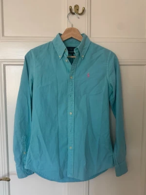Turkos skjorta från Ralph Lauren XS - Säljer en turkos långärmad skjorta från Ralph Lauren i modellen Custom Fit. Skjortan har klassisk krage, knappar framtill och en rosa broderad logga på bröstet. Perfekt för dig som gillar stilrena plagg med en färgklick.