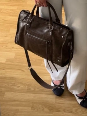 Mörkbrun handväska med axelrem - Snygg mörkbrun handväska i skinnimitation med både handtag och justerbar axelrem. Väskan har en dragkedjeficka framtill med dekorativa läderband och silvriga metalldetaljer. Perfekt storlek för att få plats med allt du behöver.