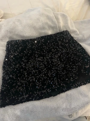 Svart paljettkjol - Säljer en svart kortkjol täckt med glittriga paljetter. Kjolen har en rak passform och är perfekt för dig som vill sticka ut med en festlig look. Materialet känns mjukt och stretchigt, vilket gör den bekväm att bära hela kvällen. Endast använd en gång