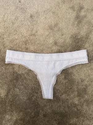 Vita stringtrosor från Victoria's Secret - Säljer ett par vita stringtrosor från Victoria's Secret med hjärtmönster och diskret spetskant. Trosorna har ett brett resårband med logotyp och är tillverkade i mjukt, stretchigt material för en bekväm passform.