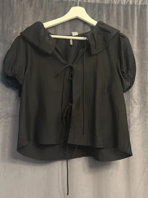 Svart blus med puffärm och knytband - Trendig svart blus med puffiga korta ärmar och dekorativ krage med spetskant. Blusen har knytband framtill och en avslappnad passform. Perfekt för dig som gillar en söt men stilren look.