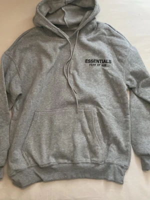 Grå Essentials Fear of God hoodie - Snygg grå hoodie från Fear of God Essentials med svart logga på bröstet och ryggen. Klassisk känguruficka framtill, huva med dragsko och mjukt material som känns riktigt skönt. Perfekt för en avslappnad streetwear.Bara testat aldrig använt