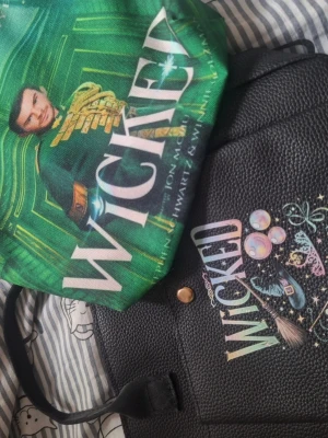 Svart handväska med Wicked-tryck - Säljer en handväska med wicked tryck och en necessär med Fiyero tigelaar från wicked. Aldrig använda 