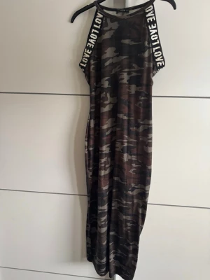 Camouflage långklänning med textband - Snygg långklänning i camouflage-mönster med svarta och bruna toner. Axelbanden har texten 'LOVE' i vitt, vilket ger en cool kontrast. Klänningen har en rak passform och är ärmlös, perfekt för en avslappnad och trendig stil.