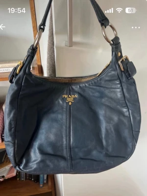 Mörkblå axelväska i läder från Prada - Snygg mörkblå axelväska från Prada i mjukt läder med guldfärgade metalldetaljer och dragkedja upptill. Väskan har en rundad form och bred axelrem med stora ringar i metall. Klassisk logga framtill i guld.