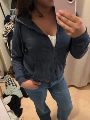 Mörkblå zip hoodie med huva - Mysig mörkblå hoodie med dragkedja och huva. Modellen har fickor framtill och ribbade muddar vid ärmslut och nederkant. Perfekt att slänga på sig över en topp eller t-shirt. Materialet ser ut att vara mjukt och skönt, troligen bomull eller bomullsblandning.
