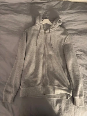 Ljusgrå zip hoodie med huva - En stilren ljusgrå hoodie med dragkedja framtill och justerbar huva med snören. Tillverkad i mjukt sweatshirtmaterial och har ribbade muddar vid ärmar och nederkant. Perfekt för en avslappnad och bekväm look.