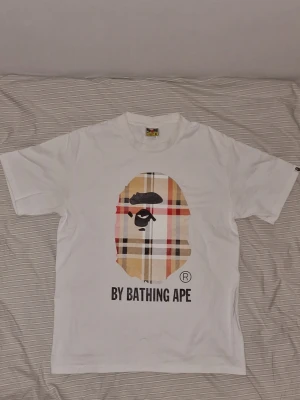 Vit BAPE t-shirt med rutigt tryck - Säljer en vit t-shirt från BAPE med det klassiska apansikte-trycket i rutig design på bröstet och texten 'BY BATHING APE' under. T-shirten har korta ärmar och är gjord i mjuk bomull. Perfekt för dig som gillar streetwear och vill sticka ut.