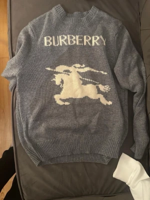 Grå stickad tröja från Burberry - Snygg grå stickad tröja från Burberry med vit logotyp och motiv av en riddare på häst på framsidan. Tröjan har rund halsringning och ribbade muddar vid ärmslut och nederkant. Perfekt för dig som gillar klassisk design med en twist.