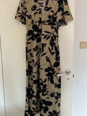 Beige/svart långklänning  - Långklänning från SHEIN i beige med stora svarta blommor. Klänningen har v-ringning, korta ärmar och en lätt utställd passform. Perfekt för dig som gillar en feminin och trendig stil.