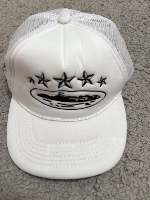 Vit truckerkeps med stjärnor - Säljer en vit truckerkeps med svart broderat motiv framtill som föreställer fem stjärnor och ett ovalt mönster. Kepsen har mesh på sidorna och justerbar snapback baktill för perfekt passform. Perfekt för dig som gillar streetwear och unik design.
