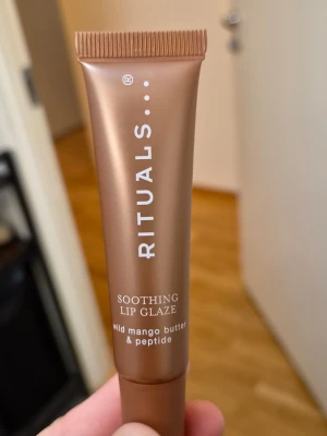 Rituals Soothing Lip Glaze - Lyxigt läppbalsam från Rituals i en snygg bronsfärgad tub. Innehåller wild mango butter och peptider som ger intensiv återfuktning och glans till läpparna. Perfekt för dig som vill ha mjuka och välvårdade läppar med en fräsch känsla.