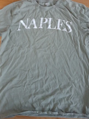 Grön t-shirt med texten NAPLES - Snygg grön t-shirt med vit text 'NAPLES' över bröstet. Klassisk passform och rund halsringning. Perfekt för en avslappnad stil och enkel att matcha med jeans eller shorts.