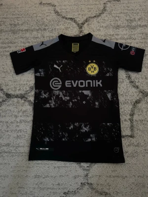 BVB Dortmund Fotbollströja - Snygg svart Borussia Dortmund matchtröja från Puma med gråa detaljer och tryck. Framsidan har klubbmärke och sponsorlogga, baksidan har Reus och nummer 11. Perfekt för fotbollsfans som vill visa sitt stöd för laget. Jätte billig och underbar fotbollströja för barn!