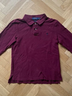 Långärmad pike Polo Ralph Lauren  - Snygg vinröd långärmad piké från Polo Ralph Lauren i slim fit-modell säljes. Tröjan är i storlek L/G 14-16 år och i mycket fint skick. 