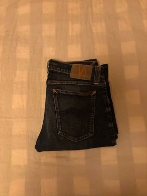 Mörkblå jeans från Nudie Jeans - Snygga mörkblå jeans från Nudie Jeans med klassisk femficksdesign och kontrastsömmar. Jeansen har en rak passform och är tillverkade i denim med en tydlig logga på bakfickan. Perfekta för en stilren och avslappnad look. Det är modellen tight long John, priset kan diskuteras