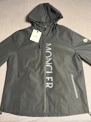 Moncler windbreaker - Snygg svart vindjacka från Moncler med stor logga vertikalt på framsidan. Jackan har huva, tre dragkedjeförsedda fickor och justerbar nederkant. Tillverkad i ett lätt och vattenavvisande material, perfekt för blåsiga dagar.