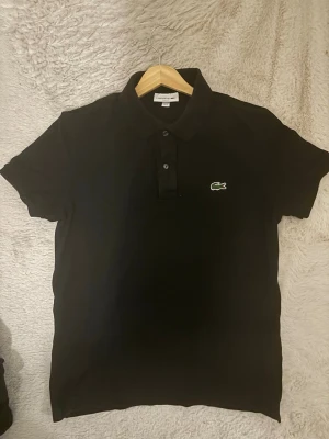 Svart pikétröja från Lacoste - Klassisk svart pikétröja från Lacoste med korta ärmar och den ikoniska gröna krokodilloggan på bröstet. Tröjan har krage och knäppning med två knappar framtill. Perfekt till både jeans och shorts.