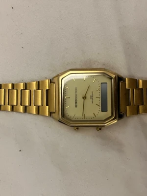 Retroinspirerad guldfärgad klocka - Snygg retroinspirerad klocka med guldfärgad länk och fyrkantig urtavla. Urtavlan är ljus med minimalistiska markeringar och digital display. Perfekt accessoar för dig som gillar vintagevibbar och stilren design.