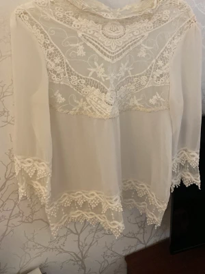 Vit spetsblus med transparenta detaljer - Säljer en romantisk vit blus med transparent tyg och vackra spetsdetaljer. Blusen har långärmad design och fina broderier längs halsringning, ärmslut och nederkant. Perfekt för dig som gillar en bohemisk och feminin stil.